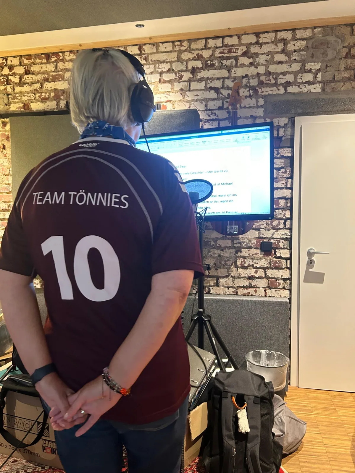 Nicki Beaujean im Team-Tönnies-Trikot mit Kopfhörern vor dem Bildschirm im Tonstudio, die Hände auf dem Rücken