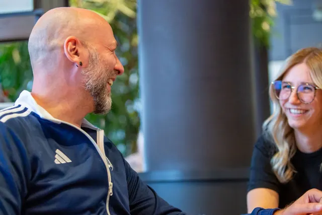Ein Mann mit Glatze und Bart in MSV-Trainingsjacke lacht, im Hintergrund lacht eine junge Frau mit Brille