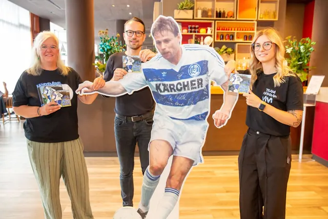 Zwei Frauen und ein Mann halten die Hörbuch-CD in die Kamera, zwischen ihnen ein lebensgroßer Aufsteller von Michael Tönnies im Kärcher-Trikot